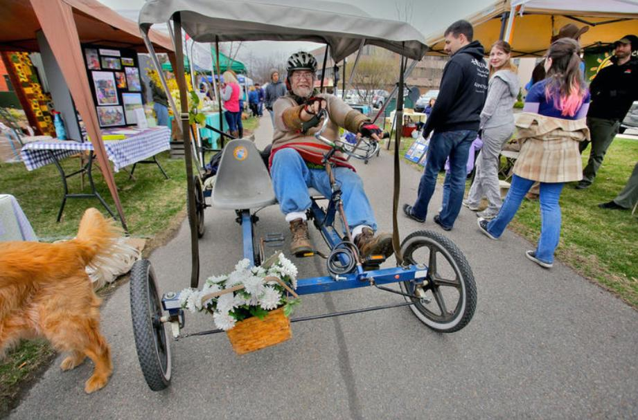 A man wearing a helmet and colorful sweater is riding a recumbent tricycle with a canopy and a flower basket on the front. He is smiling and appears to be enjoying himself at an outdoor market or fair, surrounded by booths, vendors, and people walking around. A golden retriever is visible on the left side of the image, partially out of frame. The atmosphere is lively and festive.